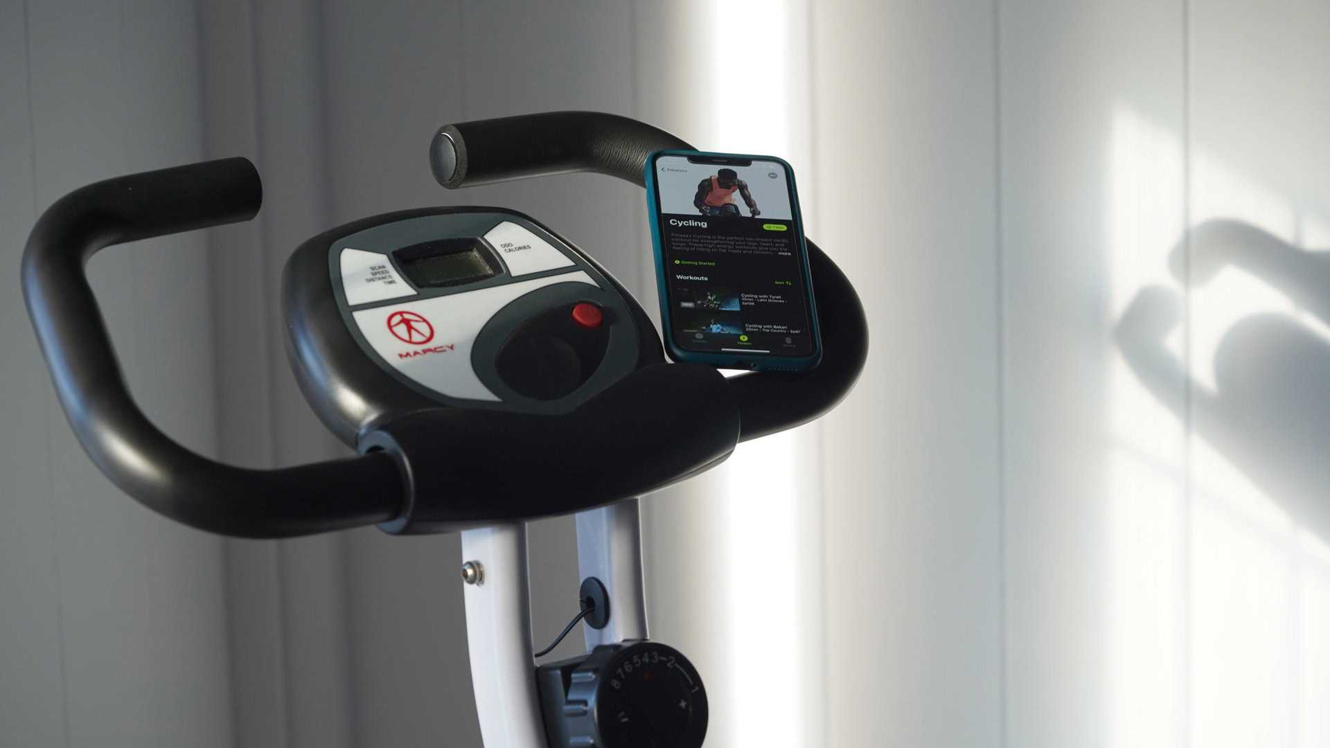 Apple Fitness Plus Cycling An iPhone resting on a stationary bike handle displays the app's Cycling menu.
